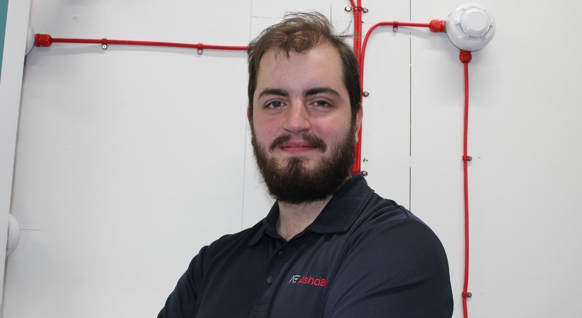 Young man with dark hair and beard wearing navy polo shirt with company name pictured in front of a fire alarm panel